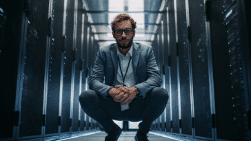 Technicien informatique moderne en costume accroupi devant une baie de serveurs, dans un centre de données sécurisé à l’ambiance high-tech.