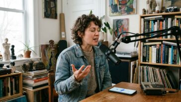 Une femme aux cheveux bouclés parle dans un microphone professionnel, assise à un bureau en bois avec un téléphone portable.